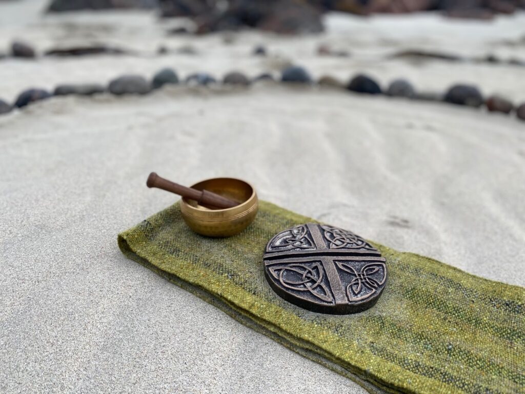 Green cloth set on the sand in a prayer circle made of rocks. A mortar and pestle and a circular metal object with Celtic symbols are set on the cloth.